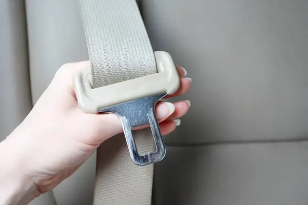 A hand holding a beige seat belt latch plate on light webbing, with the metal tongue clearly visible.